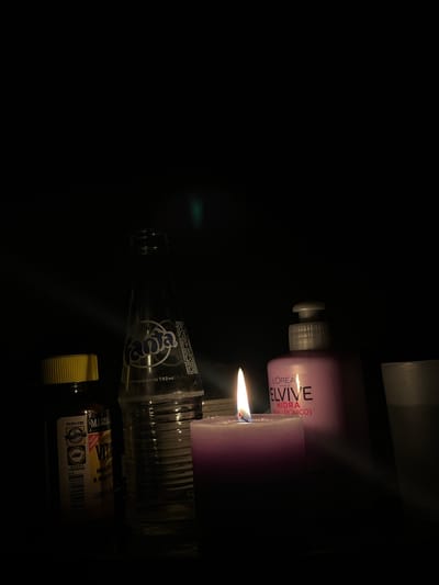 The image depicts a dimly lit scene, likely indoors, with no indication of the city of San Luis, Peru. The foreground is dominated by a lit purple candle, its flame casting a warm glow. To the left of the candle is a clear glass bottle with a Fanta label. Next to the Fanta bottle is a small brown bottle with a yellow cap, likely a supplement or medicine, and a label that includes "MASON" and "VIT." To the right of the candle is a pink bottle with a pump dispenser, labeled "L'OREAL ELVIVE." Further to the right, a portion of a light-colored glass or cup is visible. The background is entirely black, suggesting a dark room or environment. The lighting is dramatic, with rays of light from the candle illuminating the objects. There are no people or animals present in the image. The scene appears to be a still life arrangement.