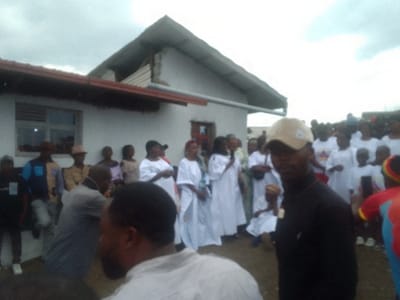 A gathering of people stands outside a white building with a gabled roof. Many are dressed in white robes. A man in a black shirt and tan baseball cap stands in the foreground, facing the camera. Other people are visible in the background wearing casual attire, and some are wearing hats. The sky appears overcast.
