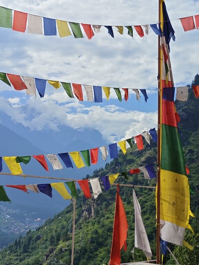 In Siumsa, India, prayer flags are strung across a mountainous landscape. Two rows of prayer flags are visible in the foreground, with a single pole holding a larger prayer flag to the right. The prayer flags are rectangular and made of fabric, displaying various colors including blue, white, red, yellow, and green. Some flags have white symbols printed on them. In the background, steep, green, forested mountains rise against a blue sky filled with white clouds. The scene depicts a clear day with good visibility. There are no people visible in the image.