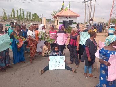 A group of women are protesting in front of the Ungwan Tanko Water Board, located at latitude: 10.422, longitude: 7.49 in Ungwan Tanko, Nigeria. Several women are holding signs with messages in English, some of which are visible. The messages on the signs include "Pay us our crops" and "All we will have from you is crumbs". The protestors are standing and sitting in the street in front of the water board building. The water board building is a small single-story structure with a brown roof and a sign on the front that reads "Ungwan Tanko Water Board". The building has a small green fence surrounding it. There are some people standing and watching the protestors from across the street. A group of men, wearing blue shirts, stand in the distance behind the protestors. The protestors are standing in a street with a paved surface and a dirt shoulder to the side. There is a green area to the left and right of the building. The sky is clear and blue. Several trees are visible in the background.