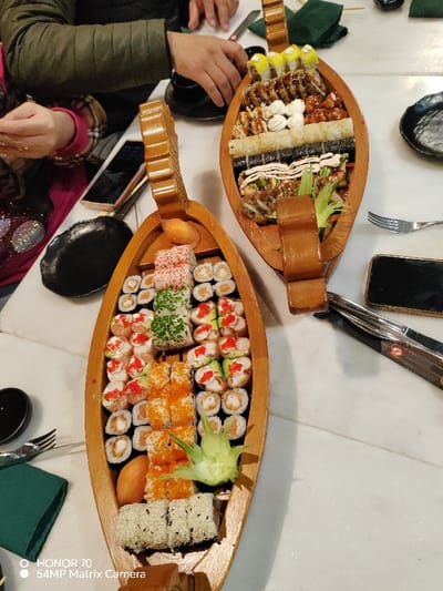 Two wooden boat-shaped platters are filled with various sushi rolls, including maki, California, and dragon rolls. One boat features rolls topped with salmon roe, avocado, and sesame seeds. The second displays unagi, tempura, and spicy tuna rolls. Chopsticks, forks, and small dishes are arranged around the platters on a white table, suggesting a shared meal setting. Two people are visible enjoying the meal.