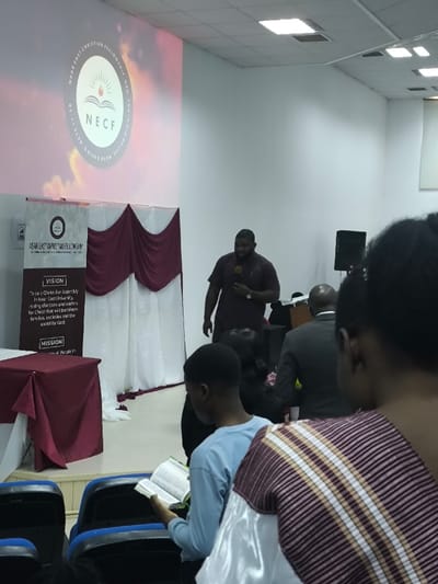 An indoor scene in Kızılbaş, Northern Cyprus / Cyprus, portrays a religious gathering or fellowship event. A male speaker, appearing to be of African descent with a beard, wears a dark short-sleeved shirt and a watch, standing centrally at the front while holding a microphone in his left hand and addressing an unseen audience. Behind him, on the left wall, a large projector screen displays a circular logo for "NEAR EAST CHRISTIAN FELLOWSHIP" (NECF) featuring an open book and radiating sun, encircled by the text "ACTS 17:28 FOR IN HIM WE LIVE AND MOVE AND HAVE OUR BEING" against a warm, cloudy background. Below the projection, a vertical banner displays the same NECF logo, name, and partially legible "VISION" and "MISSION" statements, including "To be a Christian Assembly in Near East University..." White fabric drapes with maroon swags are arranged along the wall behind the speaker. To the far left, a table is covered with a maroon tablecloth. In the immediate foreground, from a rear perspective, several audience members are seated in blue and black chairs. A young male in a light blue long-sleeved shirt is seen reading an open book, likely a Bible. Another person's back and shoulders, wearing a patterned maroon and beige striped garment over a white top, are partially visible in the extreme foreground. Further back, a male in a dark suit jacket and other individuals are partially visible, all facing the speaker. A black speaker unit rests on the floor to the speaker's right. The room is brightly lit by overhead fluorescent panel lights embedded in a white dropped ceiling.