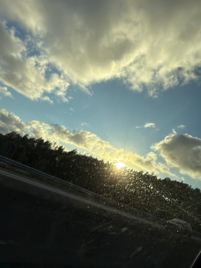 The image depicts a sky with scattered clouds, illuminated by a bright sun low on the horizon. The sun's rays are visible, creating a luminous effect against the blue sky. A dark line of trees forms the background below the sky. In the foreground, a blurry, dark surface, possibly a car windshield, is visible with numerous small, light-colored speckles. The scene suggests a view from a vehicle, possibly on a road or highway. The location in Ödsmål, Sweden, is not discernible from the visual elements of the image itself. There are no people or specific objects identified in the image beyond the natural landscape and the implied vehicle interior. The action depicted is static; it is a captured moment of scenery.