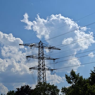 The image shows a tall, lattice-style electrical pylon silhouetted against a bright blue sky with scattered white cumulus clouds. Several power lines extend horizontally across the frame, attached to the pylon. The top of the pylon is visible, with its crossbars supporting insulators and wires. In the foreground, the tops of dark green trees are visible, partially obscuring the base of the pylon. The scene appears to be in an outdoor, possibly rural or semi-urban environment, given the presence of both the pylon and the trees. The location is identified as Forbach, Germany.