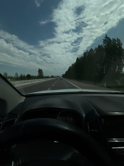 The image depicts a view from inside a vehicle, looking forward onto a highway. The road is paved with lane markings and a concrete barrier on the left. Trees line the right side of the highway, and there are scattered clouds in a blue sky. A few other vehicles are visible on the road ahead. The interior of the car, including the dashboard and steering wheel, is partially visible in the foreground. The overall impression is a travel scene on a sunny day.