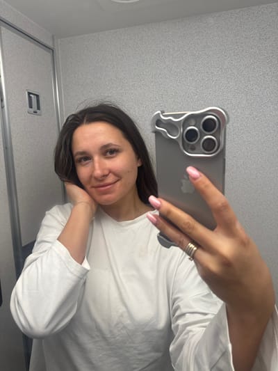 The image captures a close-up mirror selfie of a young woman, appearing to be in an enclosed space such as an airplane lavatory. The woman is smiling gently, with her head tilted slightly, and her left hand resting casually under her chin. She has long, dark, straight hair parted slightly off-center, and light-colored eyes. She is wearing a simple white, long-sleeved top. Her fingernails are manicured with light pink polish, and she wears a silver-colored ring with multiple bands on the ring finger of her right hand, and a thin silver-colored bracelet on her left wrist.
She is holding a silver-colored iPhone, likely a recent model given the large triple camera array, which is encased in a distinctive silver, possibly metallic, protective case. The case has a unique, somewhat ergonomic shape around the camera module, and the Apple logo is visible through the case. Her right hand, holding the phone, is prominently featured in the upper central part of the image.
The background, reflected in the mirror, consists of light gray, speckled, utilitarian panels on what appear to be walls and a ceiling. In the lower left portion of the image, a simple silver, horizontal door handle is visible, further suggesting a small, functional room. The lighting is even and bright, consistent with artificial indoor lighting. The overall image is rotated 90 degrees counter-clockwise from a standard portrait orientation.