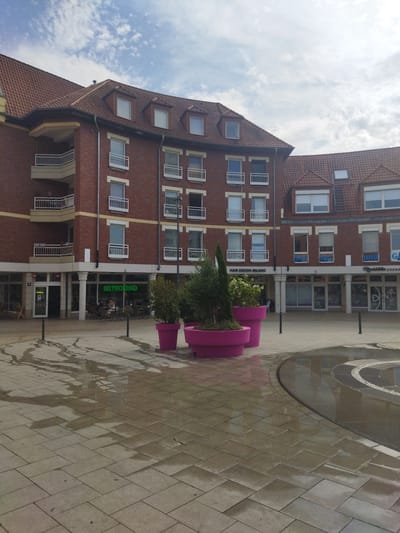 The image depicts an outdoor scene in Monheim am Rhein, Germany. In the foreground, a wet, paved courtyard reflects the sky and surrounding buildings. The paving stones are laid in a uniform pattern, with large puddles indicating recent rain or a water feature.
Dominating the background is a multi-story brick building with a red tiled roof. The architecture features several balconies with white railings on the left side and numerous windows, some with white shutters, on the upper floors. The ground level of the building appears to house several businesses, indicated by shopfronts and visible signs.
One sign reads "BISTRO 360" in bright green lettering, suggesting a restaurant or cafe. Another sign above a shop says "HAIR DESIGN MILANO." Further to the right, more shopfronts are visible, one with text that appears to include "und Kosmetik" and another with "GAM..." potentially indicating a brand or service.
In front of the shops, large, vibrant pink planters hold greenery, providing a splash of color against the muted tones of the building and paving. A few people are seated at tables outside the "BISTRO 360," suggesting a casual dining or meeting spot. The overall impression is a moderately cloudy day, likely in the afternoon, with a pleasant, albeit damp, atmosphere. The presence of businesses and outdoor seating suggests a public square or street in a commercial area.