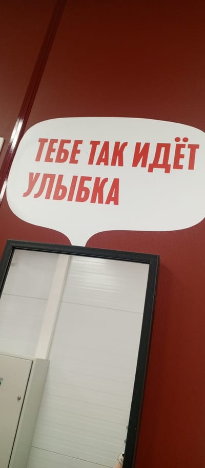 The image shows a red wall with a white speech bubble containing Russian text. The text, in red capital letters, reads "ТЕБЕ ТАК ИДЁТ УЛЫБКА". This translates to "A smile suits you so well." Below the speech bubble, a portion of a black-framed mirror is visible, reflecting a light-colored wall with horizontal lines. Part of a light-colored electrical panel with a handle is also visible in the reflection. The overall setting appears to be indoors, possibly in a public space or a business. The location is not specified in the image itself, and Zhirnovsk, Russia is not identifiable from the visual content.