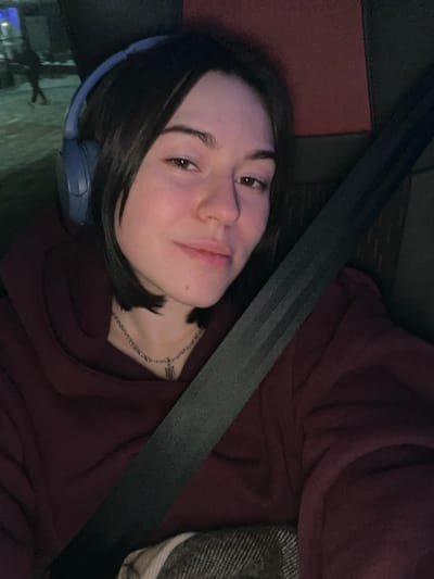 A young woman poses in a car, wearing a maroon hoodie and a seatbelt. She has short dark hair and wears headphones around her neck. A silver chain necklace is visible. The background shows a car interior with dark and red materials. Her expression is a slight smirk, angled slightly towards the camera. The overall image is somewhat dimly lit.