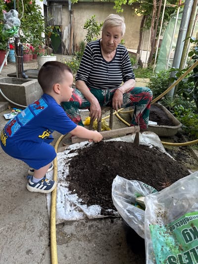 In Hadzhidimitrovo, Bulgaria, a young boy in a blue Spider-Man shirt and shorts crouches, using a small hoe to dig into a pile of dark soil. Beside him, an elderly woman with short, grey hair, wearing a striped shirt and patterned trousers, sits and observes. The scene unfolds in an outdoor setting, likely a garden or yard, with various plants, pots, and gardening equipment visible. A wheelbarrow filled with soil is present in the background, and a plastic bag with text is on the ground near the soil pile. The ground surface is a mix of concrete and earth.