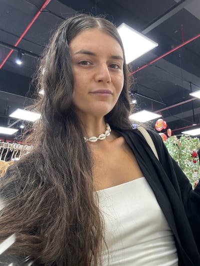 A woman with long, dark brown hair stands in a brightly lit indoor market or retail space. She is wearing a white strapless top and a black open-front cardigan. A necklace made of seashells is visible around her neck. The background shows a dark ceiling with exposed red wiring and white rectangular light fixtures. Some circular signs with red and yellow elements are visible in the upper right portion of the image. Hangers for clothing are visible to the left. The location is identified as Phu Thuy, Vietnam.