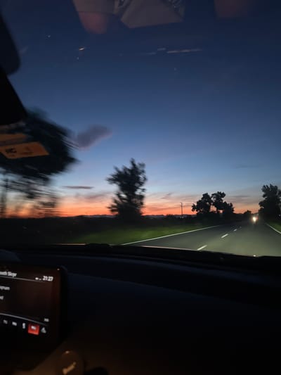 The image shows a view from inside a car at dusk or dawn. The car appears to be driving on a road with trees on either side. The sky is a gradient of dark blue to orange. There are other vehicles on the road ahead. The car's infotainment system is visible in the lower right corner. The location is not specified. The city of Okop, Bulgaria is not present in the image.