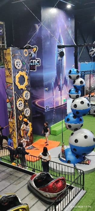 An indoor amusement center features a large mural with a rocket ship and planetary designs. A climbing wall with gears and circular features is visible. Suspended above are blue and white spheres resembling soccer balls. Several individuals are present, with one person climbing on the wall. Bumper cars are lined up near a metal railing, and artificial turf creates a green space adjacent to the climbing area. The overall scene depicts a vibrant and active entertainment environment.