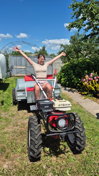 A young woman with dark hair pulled back and glasses is sitting on a small, red and black motor-cultivator with a trailer attached. She is wearing a beige crop top and pink shorts and has her arms raised in a celebratory gesture. The motor-cultivator is parked on a grassy area in front of a chain-link fence and some greenery. In the background, there are trees, a greenhouse-like structure, and a small flowering bush. The sky is blue with some scattered clouds. The location appears to be a rural area, possibly a backyard or garden in Цярэніцкі сельскі Савет, Belarus.