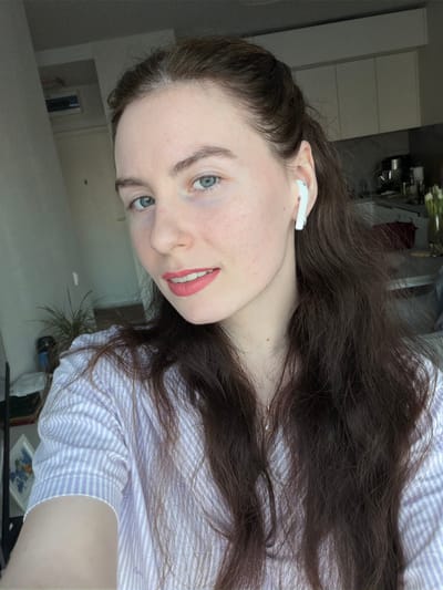 The scene is located indoors in the city of Ковалёво, Russia. A fair-skinned female, appearing to be in her mid-20s to early 30s, is positioned in the foreground, facing directly towards the camera. She has long, wavy dark brown hair, parted on the side and partially pulled back, and light blue eyes. Her lips are colored with light pink lipstick, and she exhibits a slight smile. A white wireless earbud is inserted into her right ear. She wears a light purple and white striped short-sleeved collared shirt. A delicate necklace with a small, clear or light-colored pendant is visible around her neck.
The background indicates a residential interior. To the subject's left, a light-colored wall is visible, along with a dark, upright object, possibly a door frame, and a partial view of a green potted plant. To the subject's right, a modern kitchen area is discernible, featuring white upper cabinets and a dark countertop. A vase containing several white flowers, consistent with tulips, is placed on the countertop, alongside other indistinct objects. The lighting in the scene is bright and consistent.
