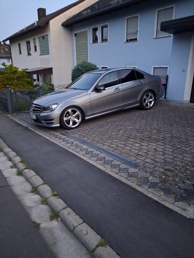 A silver Mercedes-Benz sedan is parked on a cobblestone driveway in front of a light blue and white house. The car is angled towards the right side of the image, with its front passenger side tire closest to the viewer. The house has multiple windows and a dark-colored roof. A sidewalk and a road with a curb are visible in the foreground. The scene appears to be in a residential area in Thüngersheim, Germany, given the architectural style of the houses and the presence of a license plate that includes "WÜRZBURG," a nearby major city. The lighting suggests it is either early morning or late afternoon. There are no people visible in the image.