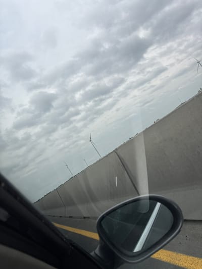 The image, shot from inside a moving vehicle, captures a scene with a prominent side mirror occupying the right side of the frame. The mirror's surface reflects the external environment. Outside the vehicle, a gray concrete barrier runs parallel to the road. Beyond the barrier, a cloudy, overcast sky fills the majority of the upper left portion of the image. Several wind turbines are visible against the cloudy backdrop. A yellow lane marking is present on the right side of the image, next to the car. The photo was likely taken near Ács, Hungary, given the presence of wind turbines in the landscape.