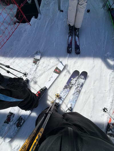 A high-angle view captures multiple individuals equipped for skiing, standing on a snow-covered surface at a ski area in Saint-Chaffrey, France. In the foreground, a person's legs are clad in black ski pants and black ski boots with red accents, holding black and yellow ski poles. To the left, another individual wears black ski pants and black boots, with a light blue strap visible on the left leg, standing on white skis featuring black and red graphics. To the mid-right, a third person is visible from the waist down, wearing white ski pants, dark ski boots, and skis with a purple and blue galaxy-like design. Partially visible on the far right are skis with green and black accents. All skis are resting on the snow, which shows tracks and disturbances. In the upper-left corner, a red safety netting and metal support structures are partially visible. Shadows cast on the snow indicate direct sunlight. The scene depicts a gathering or waiting area within a ski resort.