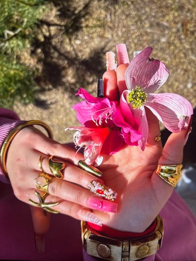 A hand holds pink dogwood and amaryllis flowers. The hand is adorned with elaborate, long fingernails, some decorated with gems and glitter. Several gold rings are on the fingers, including starfish and gem designs. A wide, patterned bone and red bracelet sits on the wrist, above a gold bangle. The blurred background shows greenery and dappled sunlight. The scene conveys a sense of nature, style, and extravagance.