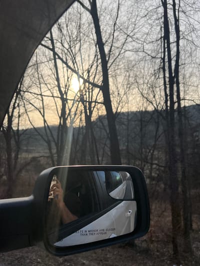 The image, captured from the interior of a vehicle in the Village of South Blooming Grove, United States of America, depicts a rural or semi-rural landscape during either sunrise or sunset. The sky, seen through a dark vehicle frame in the upper left, is bright with a prominent, low sun positioned centrally behind numerous leafless deciduous trees. These trees, characterized by their bare branches, form a dense silhouette against the sky, with a hazy line of distant hills or trees visible further back.
A black vehicle side mirror is prominently featured in the lower right quadrant. The main mirror reflects the tanned forearm and hand of a person holding a light-colored smartphone vertically, with its camera lens visible. The reflection also shows a white vehicle exterior, including a door handle, and the partial warning text "OBJECTS IN MIRROR ARE CLOSER THAN THEY APPEAR" on the lower portion of the mirror surface. A smaller, integrated convex mirror is visible in the upper left section of the main mirror, providing an expanded view of the vehicle's side.