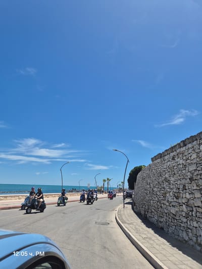 This image depicts a sunny day scene along a coastal road in Bisceglie, Italy. A group of people are riding scooters along the road, heading towards the right side of the frame. The road is paved, with a sidewalk and a low stone wall bordering the right side of the street. Beyond the sidewalk, a tall, rough-hewn stone wall extends into the distance. On the left side of the road, there is a promenade with palm trees and a view of the blue sea. The sky is clear and blue with scattered white clouds. The timestamp "12:05 ч. PM" is visible in the lower-left corner, indicating the time.
