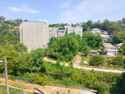 The image depicts an overcast sky with scattered clouds above a residential area in Rîbniţa, Moldova. In the foreground, a metal fence runs horizontally, bordering a dry, grassy area with a children's slide visible. Beyond the fence, a narrow body of water, possibly a stream or canal, is flanked by dense green foliage. In the mid-ground, a concrete utility pole with wires is positioned on the left side. Further back, a multi-story apartment building with a plain grey facade dominates the left half of the frame, partially obscured by lush green trees. To the right of the apartment building, a series of smaller residential houses with pitched roofs are scattered across a sloping, tree-covered hillside. A dirt road or path winds through the landscape. The overall scene suggests a mixed urban and natural environment, characterized by residential structures and abundant vegetation under a bright sky.