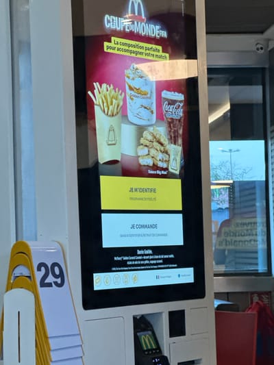 The image is a close-up of a McDonald's self-order kiosk in Narbonne, France. The screen displays an advertisement for a "Coupe du Monde FIFA" promotion, featuring french fries, a McFlurry, and Coca-Cola. Below the advertisement, there are options to "Je M'IDENTIFIE" (Identify Myself) which likely relates to a loyalty program, and "JE COMMANDE" (I Order). There is also a "Durée limitée" (Limited time) section describing the McFlurry. In the foreground, to the left, is a stack of yellow and white order number stands, with the number "29" prominently displayed. A red table or counter is visible on the right side of the image. The overall environment appears to be indoors, consistent with a fast-food restaurant.