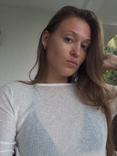 A female subject with medium-long, light brown hair, fair skin exhibiting minor blemishes, dark brown eyes, and full lips, gazes directly at the camera. She wears a light-colored, ribbed, long-sleeved top, which is semi-sheer, revealing a dark undergarment. Her left ear features two small hoop earrings. A partial dark-outlined tattoo is visible on the back of her right hand/wrist. Her right hand is positioned behind her head, partially obscured by her hair. The background consists of a plain, light-colored wall or ceiling on the left and upper left, transitioning to lush green tropical foliage, including a palm frond, on the right. The scene is set in an outdoor or semi-outdoor environment in Unawatuna, Sri Lanka, under natural light.