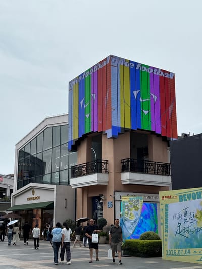 The image shows a modern street scene in Qingpu District, China, featuring commercial buildings and pedestrians. Dominating the upper portion of the image is a multi-story building with a large, colorful LED display wrapped around its corner, showcasing a "Nike football" advertisement with a rainbow of colors and Nike logos. To the left, a building with a large glass facade displays reflections of the surroundings and the text "TORY BURCH" above its entrance. Several people are walking on the paved ground in front of the stores. Some are carrying umbrellas, suggesting overcast or potentially rainy weather. One man on the right is holding a white shopping bag with "FILA" printed on it. To the right of the Nike advertisement building, another large advertisement board displays text in Chinese and English, mentioning "SUMMER" and a date "2026.07.04". The environment appears to be an outdoor shopping area or plaza.
