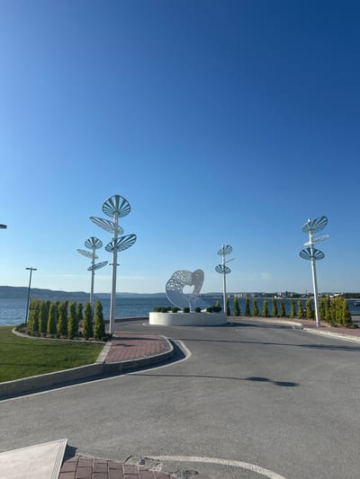 The image captures a scenic waterfront promenade on a bright, clear day, likely in the late morning or early afternoon given the strong sunlight and shadows. The scene is dominated by a vast, uninterrupted expanse of clear blue sky. In the foreground, a paved asphalt road curves along the coastline, bordered by a strip of manicured green grass and a row of small, conical evergreen trees. A patterned brick walkway runs alongside the road, adding a touch of texture and color.

Several modern, abstract sculptures, resembling stylized pinwheels or fans, are strategically placed along the promenade, mounted on tall white poles. These metallic sculptures have intricate, open designs and catch the sunlight, adding an artistic element to the outdoor space. Further along the water's edge, other decorative sculptures are visible, one appearing as a large, white, circular cutout design.

The calm, blue water of what appears to be a bay or sea stretches towards a distant hazy horizon. Across the water, a low-lying coastline with hills and buildings can be seen, suggesting a populated area. The overall atmosphere is serene and inviting, conveying a sense of leisure and public enjoyment of the waterfront. There are no people visible in the image, emphasizing the tranquil and almost deserted nature of the promenade at this moment. The clear weather and bright sunlight suggest a pleasant day for outdoor activities.