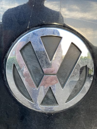 The image displays a close-up, centered view of a Volkswagen logo, likely an emblem on a vehicle. The logo is circular with a prominent chrome outline and a dark grey interior, featuring the characteristic "V" and "W" letters integrated within the circle. The chrome surface of the logo is tarnished with water spots and smudges, indicating it may be exposed to the elements. The background is a dark, glossy surface, possibly the car's paintwork, which shows a significant amount of fine scratches and some larger scuffs. A shadow, potentially from a person or object, is cast on the left side of the logo. The lighting suggests it is daytime, with natural light illuminating the scene and causing reflections on the polished surfaces. There is no visible text in the image. The context of Lütjenwestedt, Germany, places this scene in a specific geographical location but provides no direct visual cues within the image itself. The overall impression is of a somewhat weathered, but still identifiable, automotive emblem.