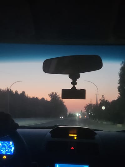The image is a dashcam view from inside a car driving on a road at dusk. The sky is a soft gradient of blue and peach, indicating sunset or sunrise. Silhouetted trees line the road on both sides, and streetlights are visible in the distance, as are car headlights. A rearview mirror with a dashcam attached is mounted to the windshield. The car's dashboard displays are visible, including an odometer reading of 166114 km and a fuel efficiency of 9.3 L/100km. The scene appears to be in or near Sariqmirzachek, Uzbekistan.