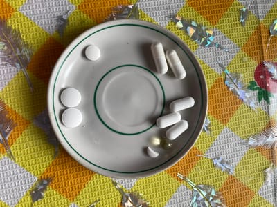 The image shows a small, white plate with a green rim, sitting on a yellow, orange, and white checkered tablecloth. On the plate are several pills: three long, white capsules; two round, white pills; one single, round, white pill; one yellowish, spherical capsule; and a small piece of a broken white pill. Decorative, shiny, feather-like objects are scattered around the plate on the tablecloth. The image does not provide information to determine the location is in Talmenka, Russia or to provide other details about people, subjects, actions or interactions.
