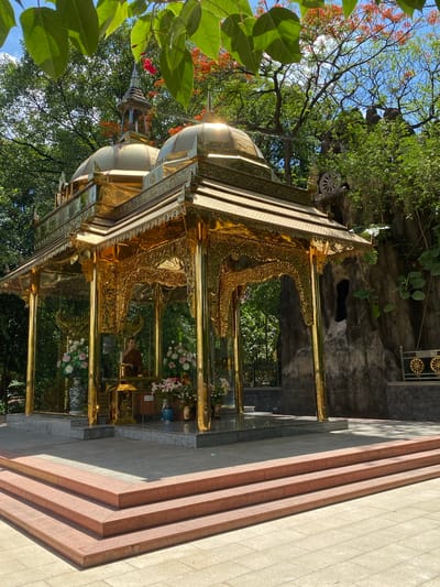 The image depicts a golden pagoda structure with a multi-tiered roof and intricate carvings, situated in an outdoor setting. The pagoda is partially obscured by foliage, with trees and flowering plants surrounding the structure. Steps lead up to the pagoda. The location is in Dĩ An, Vietnam.