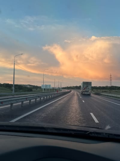 The image depicts a view from inside a moving vehicle on a highway in the Voskresensky District, Russia. The camera is facing forward, capturing the road ahead. A large truck is traveling in the same direction in front of the camera vehicle. The road is wet, and white lane markings are visible. A metal guardrail lines the side of the road. Beyond the guardrail, there is vegetation and street lights. The sky is cloudy with a sunset hue. An electrical tower is visible in the distance.