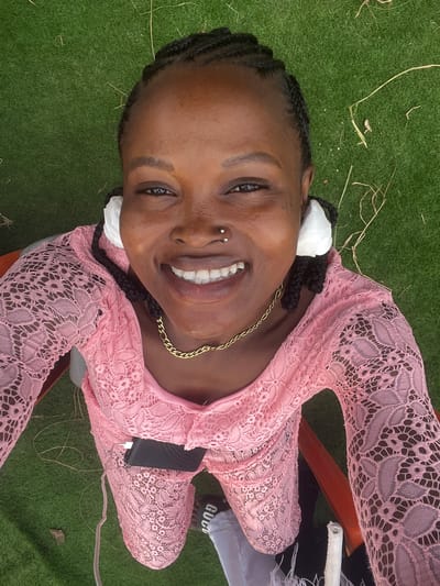 A high-angle, portrait-oriented selfie shows a young Black woman smiling broadly, with the image rotated 90 degrees clockwise as if she is lying on artificial grass. Her dark, braided hair is adorned with white, flower-like ornaments placed near her ears. She has a radiant, wide smile revealing her teeth, and multiple nose piercings: two studs on her right nostril and one on her left.

She is dressed in a light pink, long-sleeved top or dress made of delicate lace fabric with a distinct pattern. A gold-colored chain necklace is visible around her neck. A dark, rectangular object, possibly a phone or power bank, is tucked into her lap, connected by a white cable. Her right foot, wearing a sandal with the partially visible text "GUCC", is seen towards the bottom right.

The background is dominated by vibrant green artificial turf, with a few dry straw strands scattered on it. Parts of a brown or orange-colored chair are visible beneath her, suggesting she is seated. The bright, clear lighting indicates it is daytime, and her expression conveys joy and happiness. The setting is Aghalokpe, Nigeria.