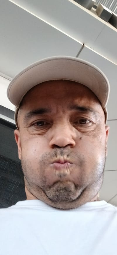 The image is a selfie taken by a man wearing a white t-shirt and a beige baseball cap. The man is looking directly at the camera with his lips pursed. He has stubble and appears to be in his 30s or 40s. The background is out of focus and appears to be indoors, possibly a modern building with clean lines and light-colored surfaces. There are no visible signs or text in the image. The lighting suggests it could be daytime. The overall mood of the image is casual and personal.