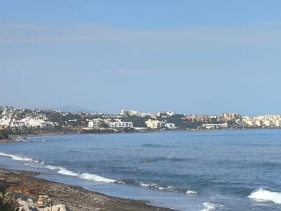 This media file depicts a scenic view of a coastline with a city nestled in the background. The foreground features a dark, pebbly beach with gentle waves lapping at the shore. In the middle ground, the calm, blue sea stretches towards the horizon, with small whitecaps indicating movement. The background is dominated by a sprawling city, characterized by numerous white buildings with red-tiled roofs, and some taller, modern structures. These buildings are situated on a hillside, backed by green trees and rolling hills, with distant mountains faintly visible under a clear, pale blue sky.

The scene appears to be set during the daytime, likely late morning or early afternoon, given the bright sunlight casting a soft illumination. The weather seems pleasant and calm, with a clear sky and tranquil waters, suggesting a peaceful coastal environment. The architecture of the buildings, with their white facades and red tiles, is characteristic of Mediterranean coastal towns. The overall impression is one of a serene and picturesque holiday destination, likely located in a warm climate. No people or specific activities are evident in the image, emphasizing the natural beauty of the landscape and the urban development along the coast. The location is identified as La Gaspara, Spain, which aligns with the visual cues of Mediterranean architecture and coastal scenery. There is no visible text in the media file.