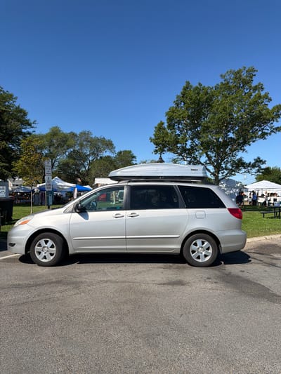 An outdoor scene in the Village of Sag Harbor, United States of America, features a silver minivan parked on asphalt. The minivan has a silver cargo box mounted on its roof and is positioned to the right of the frame. In the background, a grassy area is dotted with white tents, suggesting a market or outdoor event. Trees with green foliage are visible behind the tents, and a clear blue sky dominates the upper portion of the image. Several signs are posted near the minivan, including one that reads "NO PARKING FOR TOW AWAY ZONE" and another that says "NO DOGS ALLOWED IN PARK." The lighting indicates a bright, sunny day.