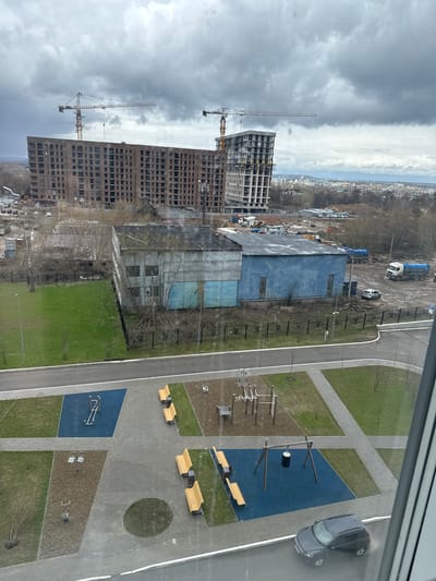 This high-angle, panoramic view from a building overlooks an urban landscape in Matiushino, Russia, under an overcast sky.

In the immediate foreground, a modern recreational area is visible, featuring paved walkways, patches of green grass, and designated play or exercise zones with blue rubberized surfaces and sand-colored ground cover. These zones contain various equipment, including monkey bars, a small balance beam, and other fitness structures. Several light-colored wooden benches are placed along the paved paths. A dark grey car is parked on a paved road bordering this area.

Further into the midground, a large, rectangular industrial-style building with blue siding stands. Adjacent to it is an active construction site with several large trucks, including cement mixers, and piles of earth or materials. Scattered bare trees indicate late winter or early spring. A blue "P" sign with an arrow, indicating parking, is visible on a post near the construction vehicles.

The background features multiple tall, multi-story buildings. Some appear to be completed residential structures with numerous windows, while others are clearly under construction, showing concrete frameworks and scaffolding. Several prominent yellow tower cranes are visible, reaching high into the heavy, dark grey cloud-filled sky. A distant cityscape or town is discernible on the horizon, blending with the cloudy weather conditions.

No people are visible engaged in activities within the scene. The overall impression is one of ongoing urban development, contrasting newly completed public spaces with active construction and industrial elements.