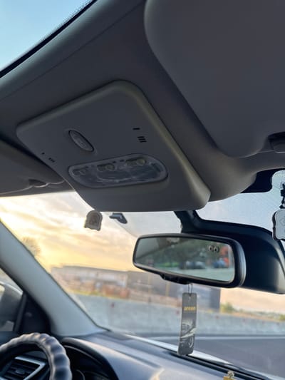 The image shows the interior of a vehicle, looking forward through the windshield. The sky is visible with clouds and a sunset. In the foreground, a steering wheel is partially visible. Above the windshield, there is a console with buttons and lights, and a rearview mirror. A car air freshener hangs from the rearview mirror. The background appears to be a highway with some buildings. There are no people visible in the image. The location is identified as Cojușna, Moldova.