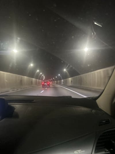 This media file captures a view from inside a vehicle traveling through a tunnel in Dolna Vasilitsa, Bulgaria. The scene is dark, illuminated by the tunnel's lighting system, which consists of a series of lights running along the ceiling and walls. The tunnel appears to be a road tunnel, with a lane visible ahead leading into the distance. A red vehicle with illuminated taillights is visible further down the tunnel, suggesting other traffic is present.
The perspective suggests the photo was taken from the driver's or passenger's seat, looking forward through the windshield and side window. Part of the vehicle's interior, including the dashboard and a vent, is visible on the right side of the frame, indicating the point of origin of the photograph. The tunnel's architecture is somewhat industrial, with concrete walls and ceiling. The time of day is indeterminate due to the enclosed nature of the tunnel, but the darkness outside and the artificial lighting suggest it could be nighttime or during overcast conditions. There is no explicit mention of weather conditions, but the overall atmosphere is somewhat damp or misty, potentially due to the enclosed environment. No people are clearly visible. A faint logo or text, possibly "GT", is visible on the dashboard, but it is not clear enough to read definitively. The overall impression is one of forward motion and passage through an enclosed, artificial space.
