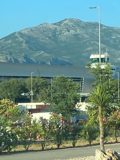 The image depicts a view of the Dubrovnik Airport (DBV) control tower and terminal building, situated in Močići, Croatia. In the foreground, flowering oleander bushes in shades of pink and white line a paved walkway. Behind the bushes, a modern airport terminal building with a dark, sloping roof is visible, and to its right, the airport control tower stands tall. The control tower is a white structure with green-tinted windows and a white light pole extending upwards next to it. In the background, a large, rugged mountain range under a clear blue sky dominates the horizon. The overall scene is one of an airport infrastructure integrated with a natural, mountainous landscape.