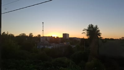 The image captures a view of Volkhov, Russia, during sunset. The sky is a gradient of pale blue to soft orange and yellow as the sun dips below the horizon, partially obscured by trees and buildings. In the foreground, dense green foliage dominates the lower half of the frame, with some taller trees casting shadows. In the middle ground, a collection of low-rise industrial or utilitarian buildings are visible, some with visible signs of age. To the left, a tall, lattice-style radio or communication tower stands prominently against the sky, reaching above the treeline. Further to the right, a multi-story residential building with numerous windows is visible. The overall impression is an urban landscape at dusk, with elements of industry, residential life, and natural greenery. There are no people or animals visible in the frame.