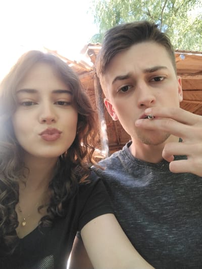 This is a selfie taken outdoors, likely during the day given the bright lighting. Two young people are visible: a woman on the left and a man on the right. The woman has long, curly brown hair and is looking at the camera with her lips pursed as if for a kiss. She is wearing a black t-shirt and a delicate gold necklace with a heart pendant. The man, with short brown hair, is also looking towards the camera, and he is holding a lit cigarette to his lips. He is wearing a dark gray, textured t-shirt.

Behind them, a wooden structure with a thatched or corrugated roof is visible, suggesting an outdoor seating area or shelter. Lush green foliage from trees is also present in the background, further indicating an outdoor setting, possibly in a rural or park-like environment. The overall mood appears casual and relaxed. There is no visible text in the image. The provided location context is Miločani, Montenegro.