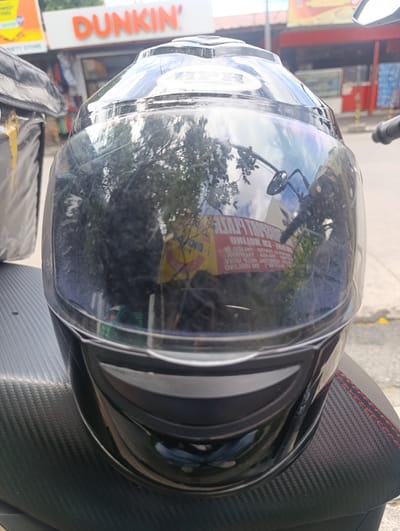 A black motorcycle helmet with a reflective visor is positioned centrally in the foreground, resting on a black seat with red stitching. The helmet's visor reflects trees, sky, and a building with the "Dunkin'" logo, indicating a location in Imus, Philippines. The background shows a Dunkin' establishment and a street with some pedestrians visible. The overall scene is outdoors, under daylight conditions.