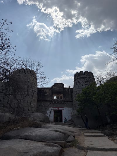 The image depicts the ruins of the Chitradurga Fort, located in the city of Chikkarampur, India, against a backdrop of a partly cloudy sky with sun rays. The ruins feature stone structures with cylindrical turrets, weathered stonework, and arched openings. The foreground includes rocky terrain and sparse vegetation. There are 2 indistinct people in the background.