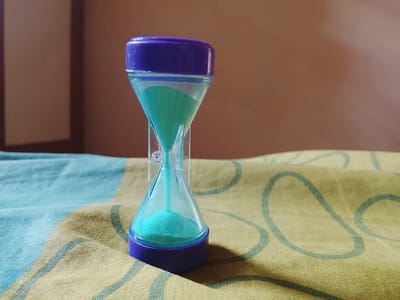 An hourglass with purple caps and clear plastic casing is positioned on a patterned fabric surface. The hourglass contains turquoise sand, with a portion of the sand accumulated in the bottom bulb and a stream of sand falling from the top bulb. The background consists of a brown, out-of-focus wall with a vertical shadow. The fabric beneath the hourglass displays a pattern of beige and muted blue stripes, overlaid with light green circular designs. The scene is lit from the side, casting a shadow from the hourglass onto the fabric. The image is taken from a slightly elevated angle. The described scene is not specific to Karangklesem, Indonesia, and lacks any contextual elements to place it within that location.