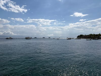 The image captures a wide-angle view of a body of water, likely the ocean, with several boats of various sizes dotting the surface. In the distance, a lush green coastline is visible, dotted with buildings and palm trees, suggesting a tropical location. The sky is a vibrant blue, partially filled with scattered white clouds. The water itself is a deep blue, with sunlight glinting off its surface, creating a shimmering effect. The scene appears peaceful and serene, likely depicting a coastal area or harbor. The location is identified as Kampung Toyapakeh, Indonesia, known for its beautiful beaches and waters.
