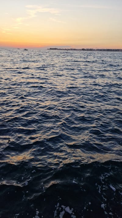 This media file depicts a sunset over the Adriatic Sea, viewed from the coast of Zadar, Croatia. The scene is split horizontally: the left side features a soft gradient of orange and yellow hues in the sky, transitioning to a lighter yellow near the horizon, indicative of the setting sun. Scattered clouds are visible in the sky. The right side of the image is dominated by the dark, choppy surface of the sea, with the texture of small waves and white foam.

The horizon line is visible in the distance, separating the sky and sea. Along the horizon, a subtle silhouette of a city skyline can be discerned, with buildings and perhaps some masts from boats. A small boat is also visible in the water, closer to the horizon. The colors of the sunset cast a warm, golden light onto the crests of the waves, contrasting with the deep blue and dark tones of the water. The overall mood is serene and peaceful, characteristic of a sunset by the sea. The time of day is clearly sunset, and the weather appears calm with gentle waves. There is no visible text in the image.